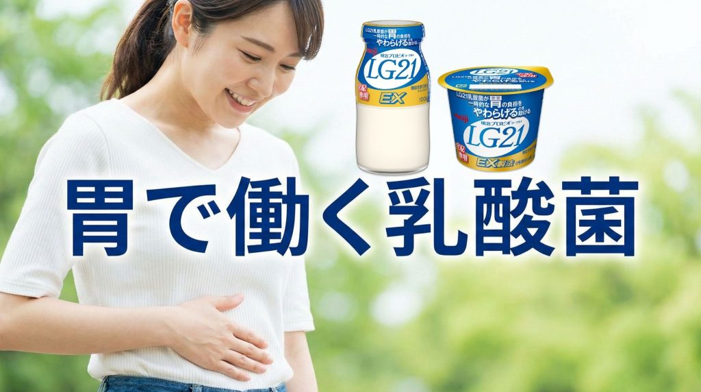 屋外の自然な背景で、女性がお腹に手を当てて微笑む、横長の写真。「胃で働く乳酸菌」の文字が大きく配置されている。